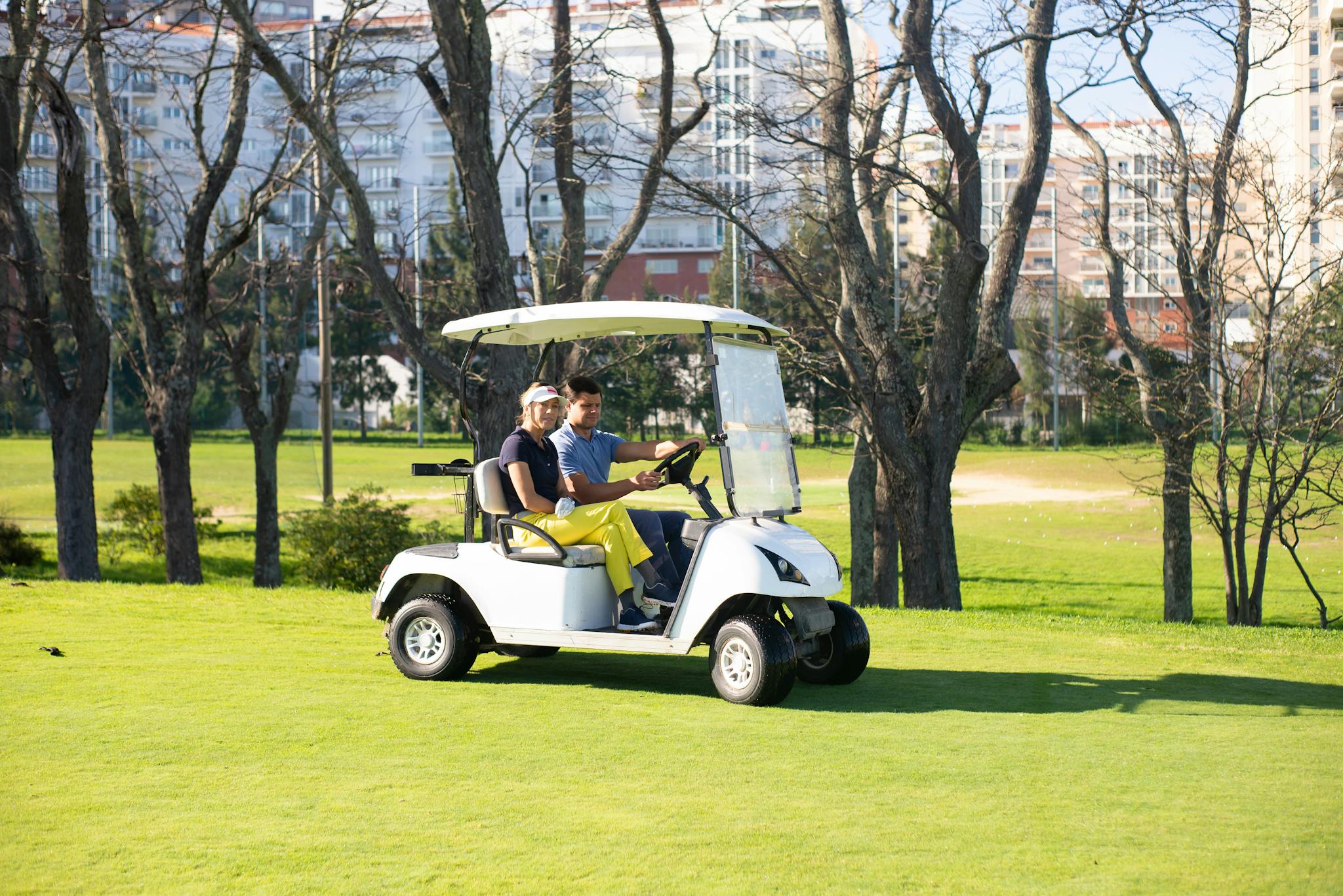 A couple rides a golf cart in a sunny park with green grass and trees.